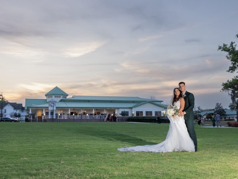 A bride and groom standing with Ocoee Lakeshore Center in the background on a grass field.