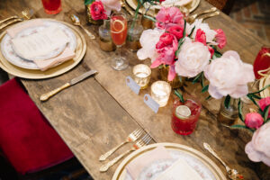 A wood table with gold rim chargers, gold flatware, pink napkins as well as pink and white florals as centerpieces.